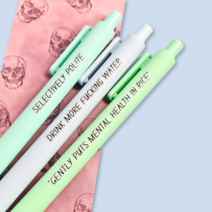 Three pens with humorous text on a light blue background. Text reads, "Gently puts mental health in rice" on a pastel green pen, "Selectively polite" on a mint pen and "drink more fucking water" on a pastel blue pen
