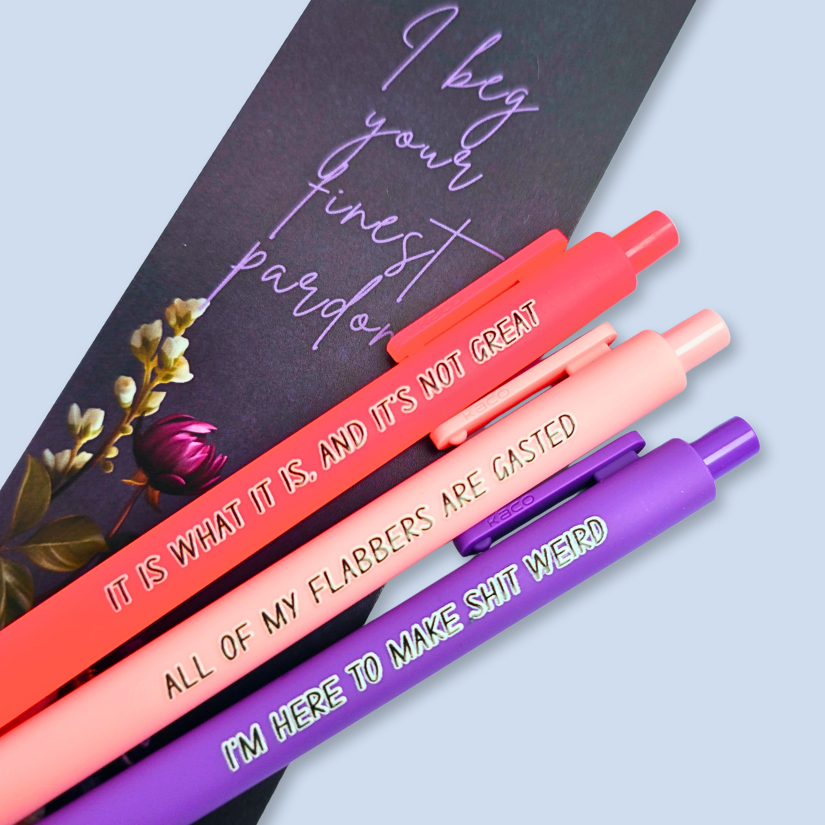 Three pens with sayings on them. "All of my flabbers are gasted" (pastel pink pen), "I'm here to make shit weird" (purple pen), "It Is what it is, and it's not great" ( neon pink pen).