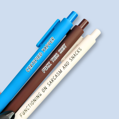 Three pens with sayings on them. "certified yapper" (bright blue pen), "Functioning on sarcasm and snacks" (white pen), "fuck this shit" (brown pen).