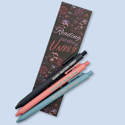 Three pens with quotes. "Reading something Unholy" on a navy blue pen, "smut reader. Probably feral." on a pink pen, and "burn my Kindle when I die" on a grey blue pen. Bookmark in the background  has a floral and skulls pattern