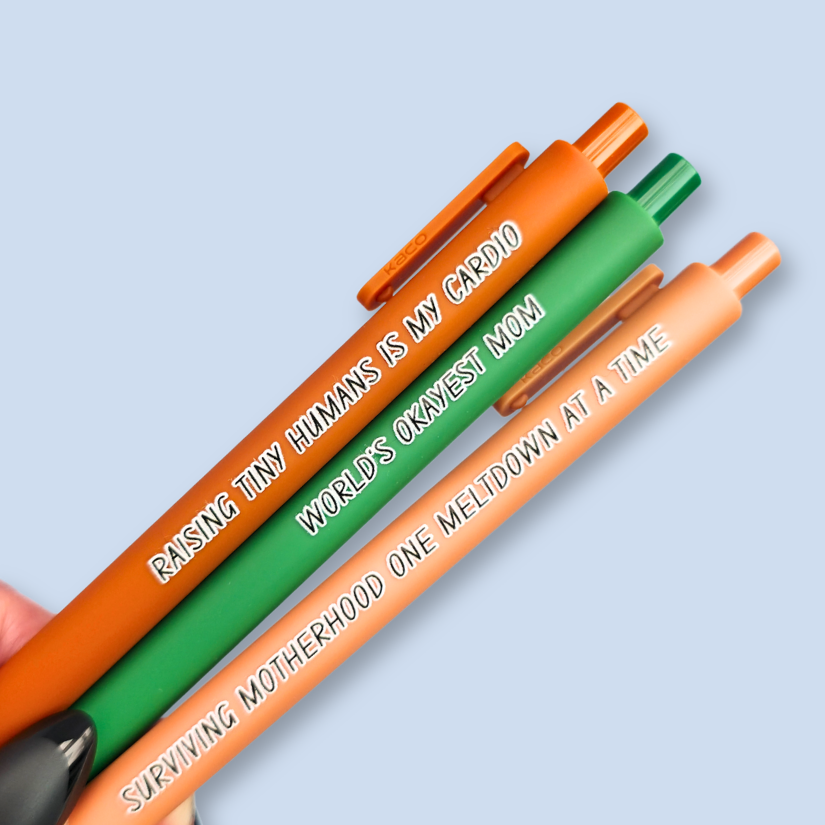 Three pens with humorous text on a light blue background. Text reads, "Raising tiny humans is my cardio" on a sienna pen, "World's okayest mom" on a green pen and "surviving motherhood one meltdown at a time" on a caramel pen