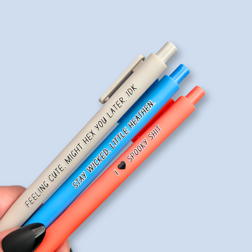 Three pens with humorous text on a light blue background. Text reads, "Feeling cute, might hex you later, IDK" on a light grey pen, "Stay wicked, little heathen" on a bright blue pen and "I  heart spooky shit" on a orange pen