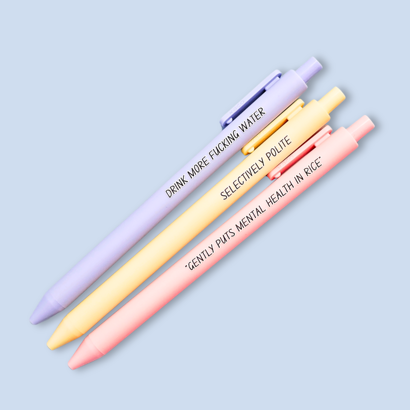 Three pens with humorous text on a light blue background. Text reads, "Gently puts mental health in rice" on a pastel pink pen, "Selectively polite" on a pastel yellow pen and "drink more fucking water" on a lavender pen