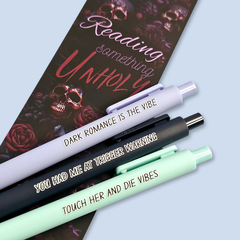 Three pens with sayings on them. "Dark romance is the vibe" (lavender pen), "You had me at trigger warning" (black pen), "Touch her and die vibes" ( mint pen).