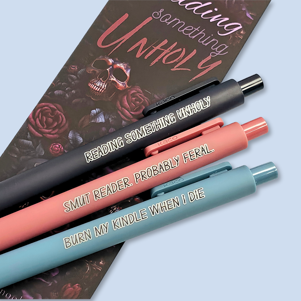 Three pens with quotes. "Reading something Unholy" on a navy blue pen, "smut reader. Probably feral." on a pink pen, and "burn my Kindle when I die" on a grey blue pen.