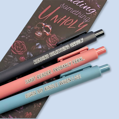 Three pens with quotes. "Reading something Unholy" on a navy blue pen, "smut reader. Probably feral." on a pink pen, and "burn my Kindle when I die" on a grey blue pen.