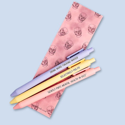 Three pens with humorous text on a light blue background. Text reads, "Gently puts mental health in rice" on a pastel pink pen, "Selectively polite" on a pastel yellow pen and "drink more fucking water" on a lavender pen
