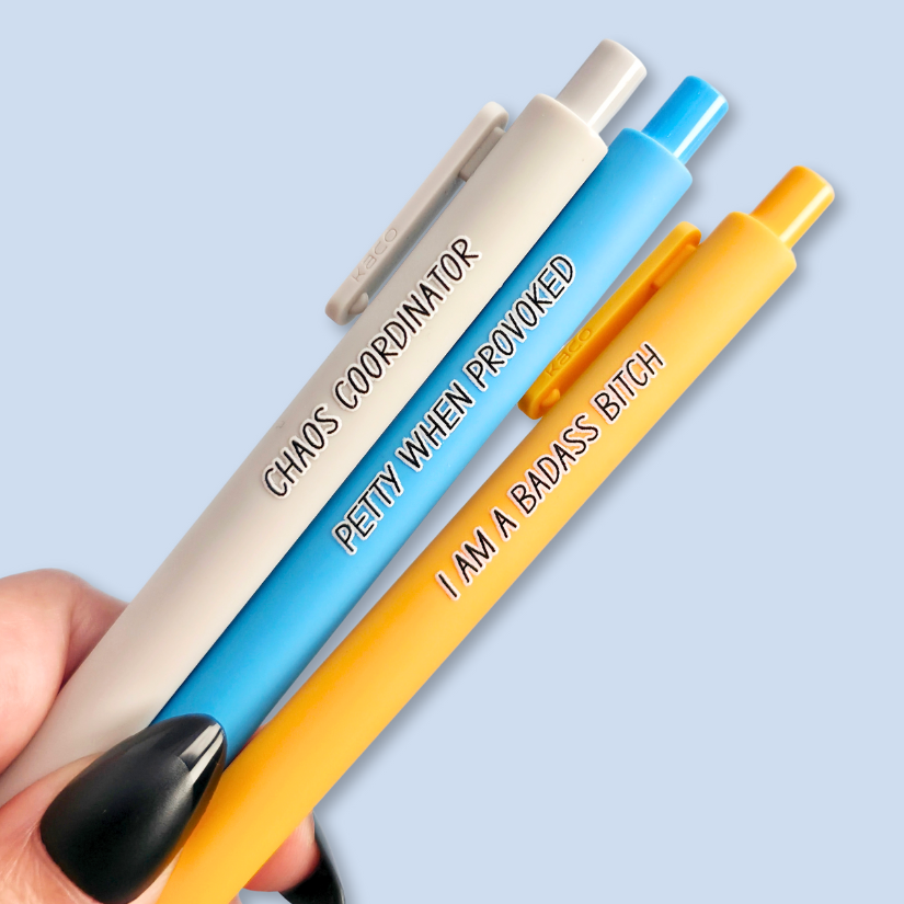 Three pens with humorous text. "Petty when provoked" on a bright blue pen, "chaos coordinator" on a light grey pen, and "I am a badass bitch" on a yellow pen