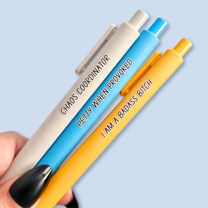 Three pens with humorous text. "Petty when provoked" on a bright blue pen, "chaos coordinator" on a light grey pen, and "I am a badass bitch" on a yellow pen