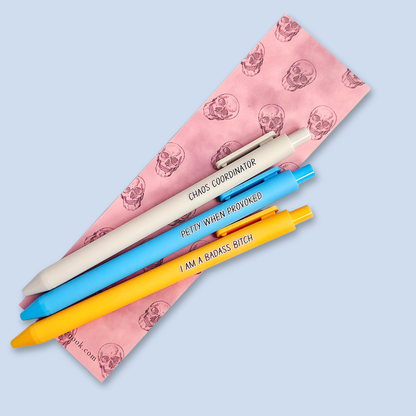 Three pens with humorous text. "Petty when provoked" on a bright blue pen, "chaos coordinator" on a light grey pen, and "I am a badass bitch" on a yellow pen