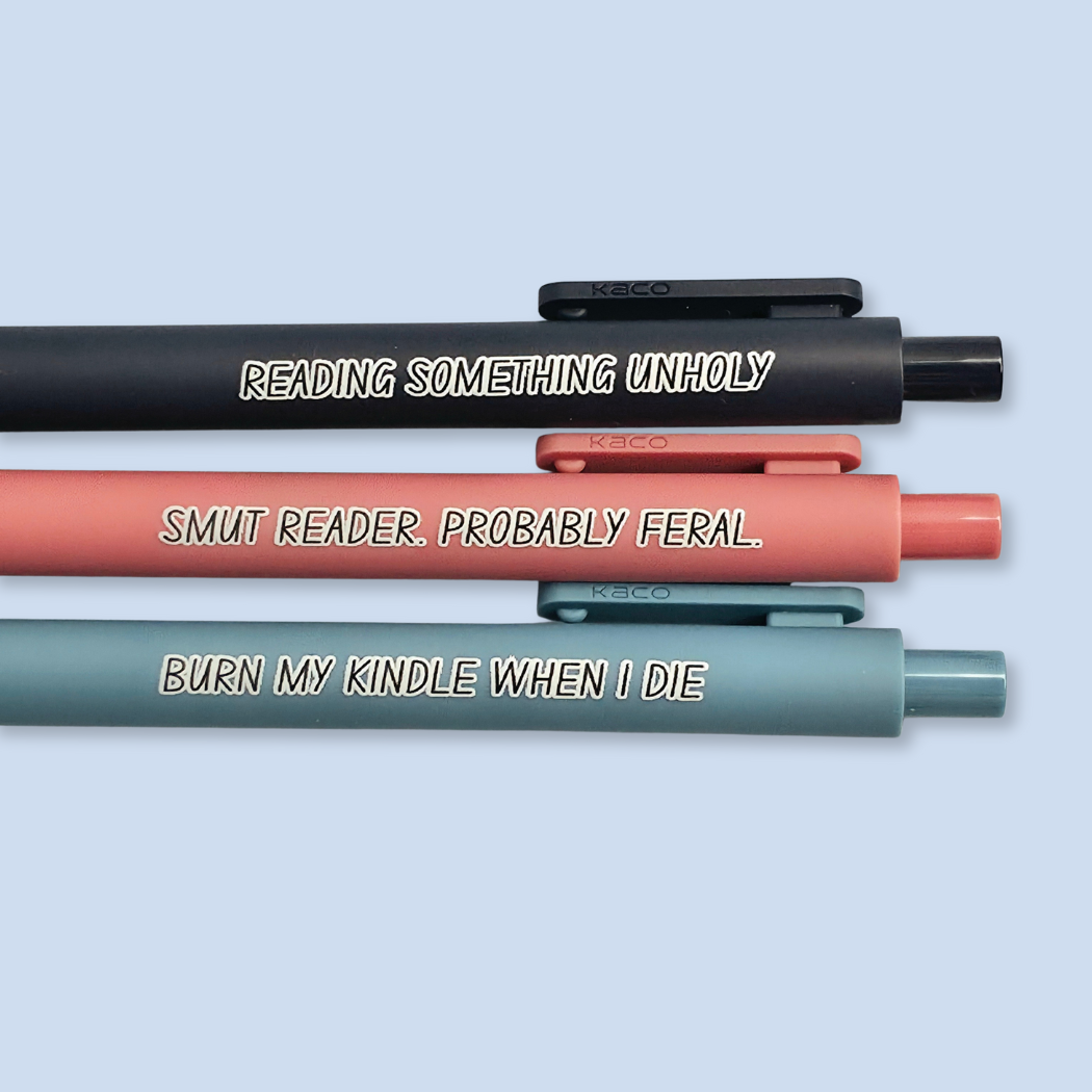Three pens with quotes. "Reading something Unholy" on a navy blue pen, "smut reader. Probably feral." on a pink pen, and "burn my Kindle when I die" on a grey blue pen.