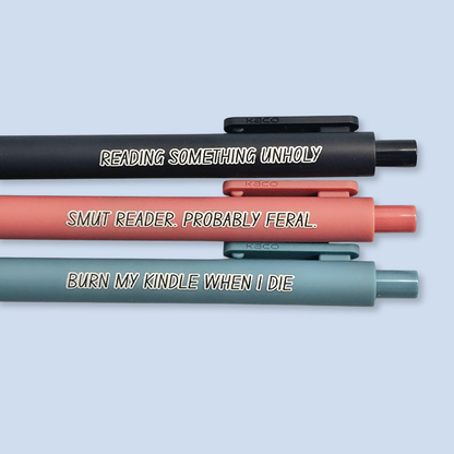 Three pens with quotes. "Reading something Unholy" on a navy blue pen, "smut reader. Probably feral." on a pink pen, and "burn my Kindle when I die" on a grey blue pen.