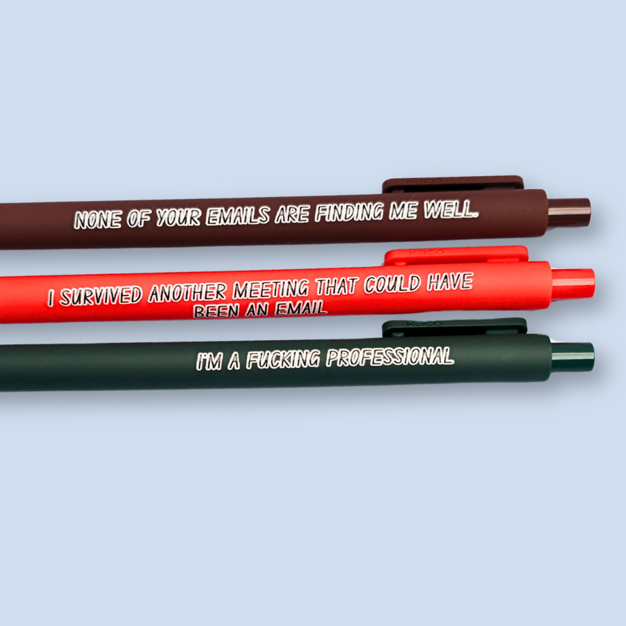 Three pens with quotes. "None of your emails are finding me well" on a brown pen, " I'm a fucking professional" on a forest green pen, and "I survived another meeting that could have been an email" on a orange pen.