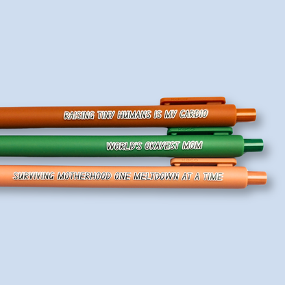 Three pens with humorous text on a light blue background. Text reads, "Raising tiny humans is my cardio" on a sienna pen, "World's okayest mom" on a green pen and "surviving motherhood one meltdown at a time" on a caramel pen