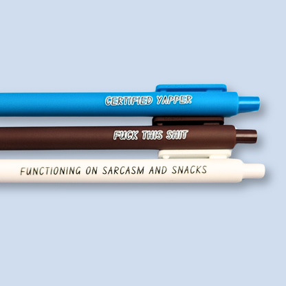Three pens with sayings on them. "certified yapper" (bright blue pen), "Functioning on sarcasm and snacks" (white pen), "fuck this shit" (brown pen).