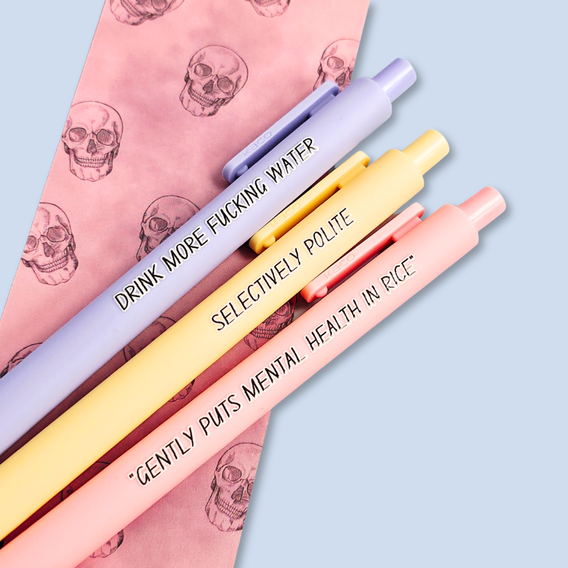 Three pens with humorous text on a light blue background. Text reads, "Gently puts mental health in rice" on a pastel pink pen, "Selectively polite" on a pastel yellow pen and "drink more fucking water" on a lavender pen