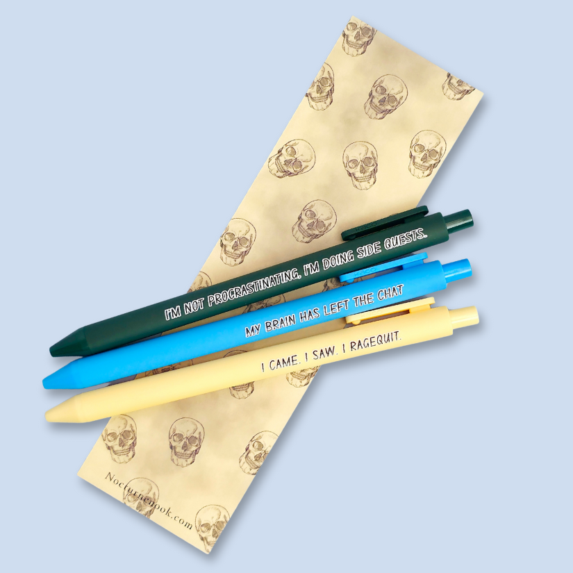 Three pens with humorous text on a skull-patterned bookmark against a light blue background