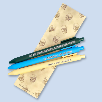 Three pens with humorous text on a skull-patterned bookmark against a light blue background