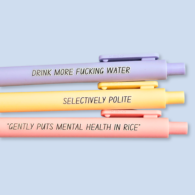 Three pens with humorous text on a light blue background. Text reads, "Gently puts mental health in rice" on a pastel pink pen, "Selectively polite" on a pastel yellow pen and "drink more fucking water" on a lavender pen