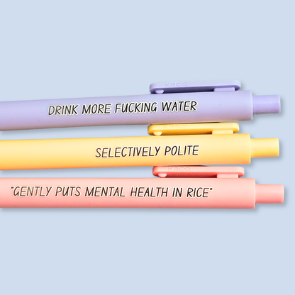 Three pens with humorous text on a light blue background. Text reads, "Gently puts mental health in rice" on a pastel pink pen, "Selectively polite" on a pastel yellow pen and "drink more fucking water" on a lavender pen
