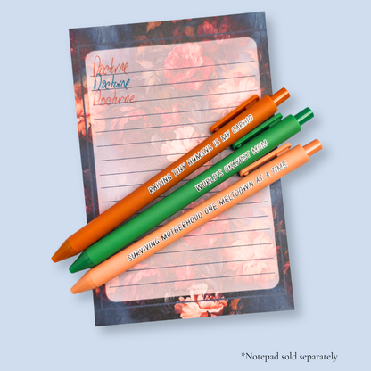 Three pens with humorous text on a light blue background. Text reads, "Raising tiny humans is my cardio" on a sienna pen, "World's okayest mom" on a green pen and "surviving motherhood one meltdown at a time" on a caramel pen. Ink matches the color of the pen