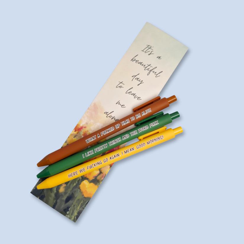 Three pens with sayings on them. "here we fucking go again. I mean, Good morning!"(bright yellow pen), "What a fucked up time to be alive" (sienna pen), "I like pretty things and the word fuck" ( green pen) bookmark on background
