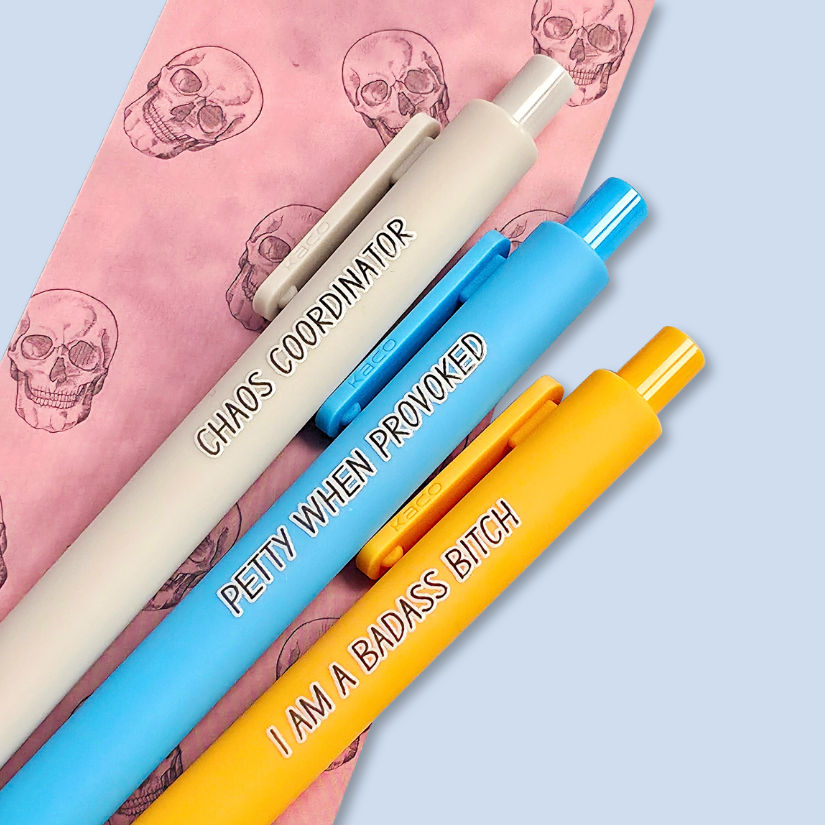 Three pens with humorous text. "Petty when provoked" on a bright blue pen, "chaos coordinator" on a light grey pen, and "I am a badass bitch" on a yellow pen
