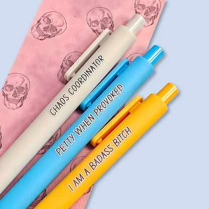 Three pens with humorous text. "Petty when provoked" on a bright blue pen, "chaos coordinator" on a light grey pen, and "I am a badass bitch" on a yellow pen