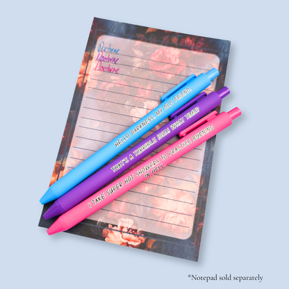 Three pens with quotes. "Hello darkness, my old friend." on a bright blue pen, ' That's a terrible idea. What time?" on a purple pen, and "I take super hot showers to practice burning in hell" on ahot pink pen. The pens rest on a notepad, with a sample of the ink colors. Notepads are sold separately