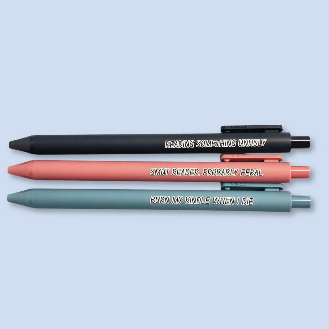 Three pens with quotes. "Reading something Unholy" on a navy blue pen, "smut reader. Probably feral." on a pink pen, and "burn my Kindle when I die" on a grey blue pen.