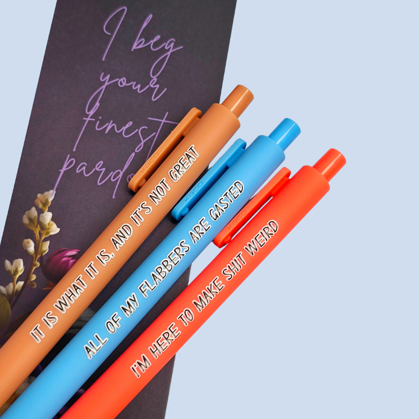 Three pens with sayings on them. "All of my flabbers are gasted" (bright blue pen), "I'm here to make shit weird" (neon orange pen), "It Is what it is, and it's not great" ( caramel pen).