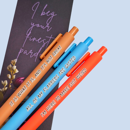 Three pens with sayings on them. "All of my flabbers are gasted" (bright blue pen), "I'm here to make shit weird" (neon orange pen), "It Is what it is, and it's not great" ( caramel pen).