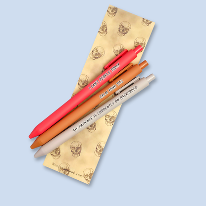 Three pens on a yellow bookmark with skull designs on a light blue background