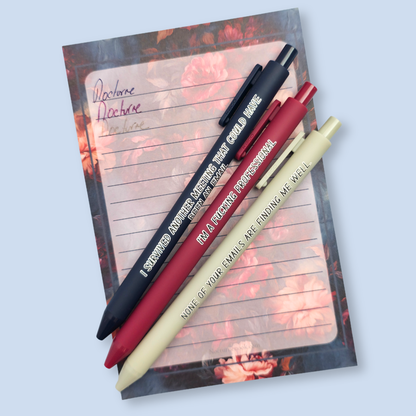 Three pens with quotes. "None of your emails are finding me well" on a grey pen, " I'm a fucking professional" on a dark red pen, and "I survived another meeting that could have been an email" on a navy blue pen.