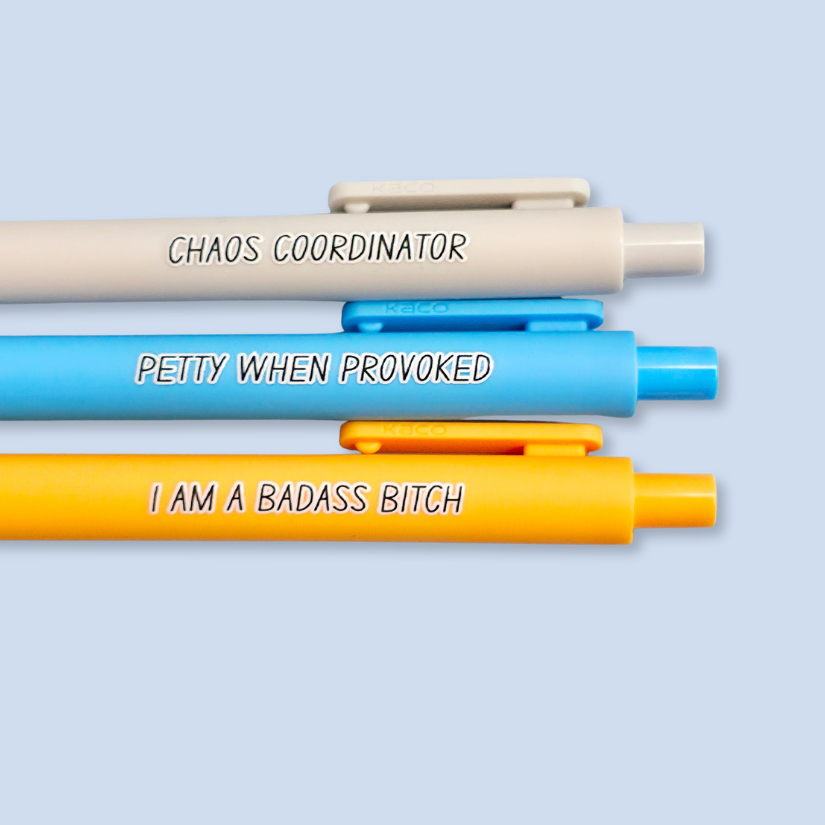 Three pens with humorous text. "Petty when provoked" on a bright blue pen, "chaos coordinator" on a light grey pen, and "I am a badass bitch" on a yellow pen