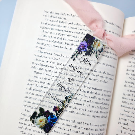 Decorative floral bookmark on an open book with visible text. The bookmark reads "You had me at trigger warning" and has a floral design. Colors of flowers are blue, purple, and white with green foliage. 