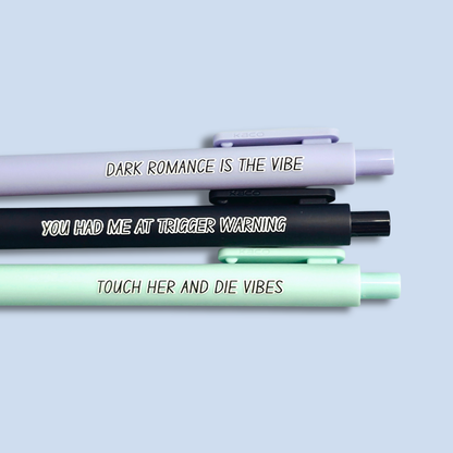 Three pens with sayings on them. "Dark romance is the vibe" (lavender pen), "You had me at trigger warning" (black pen), "Touch her and die vibes" ( mint pen).