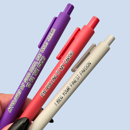 Three novelty gel pens in purple, coral pink, and light grey, held in a hand against a light blue background. Each pen features humorous printed text on the barrel.